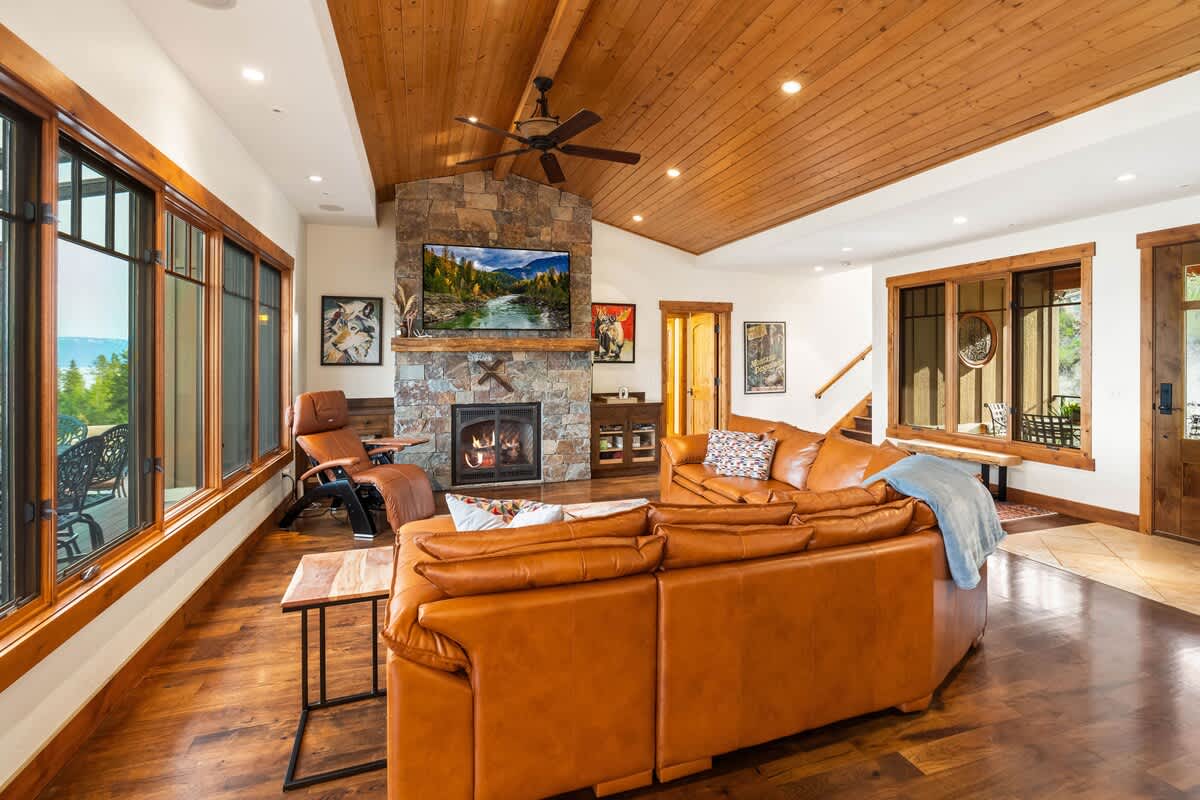 A cozy living room centered around a large brown leather sectional sofa and a prominent stone fireplace with a TV above. The room boasts a vaulted wood-paneled ceiling, hardwood floors, and multiple windows providing views of the surrounding nature.