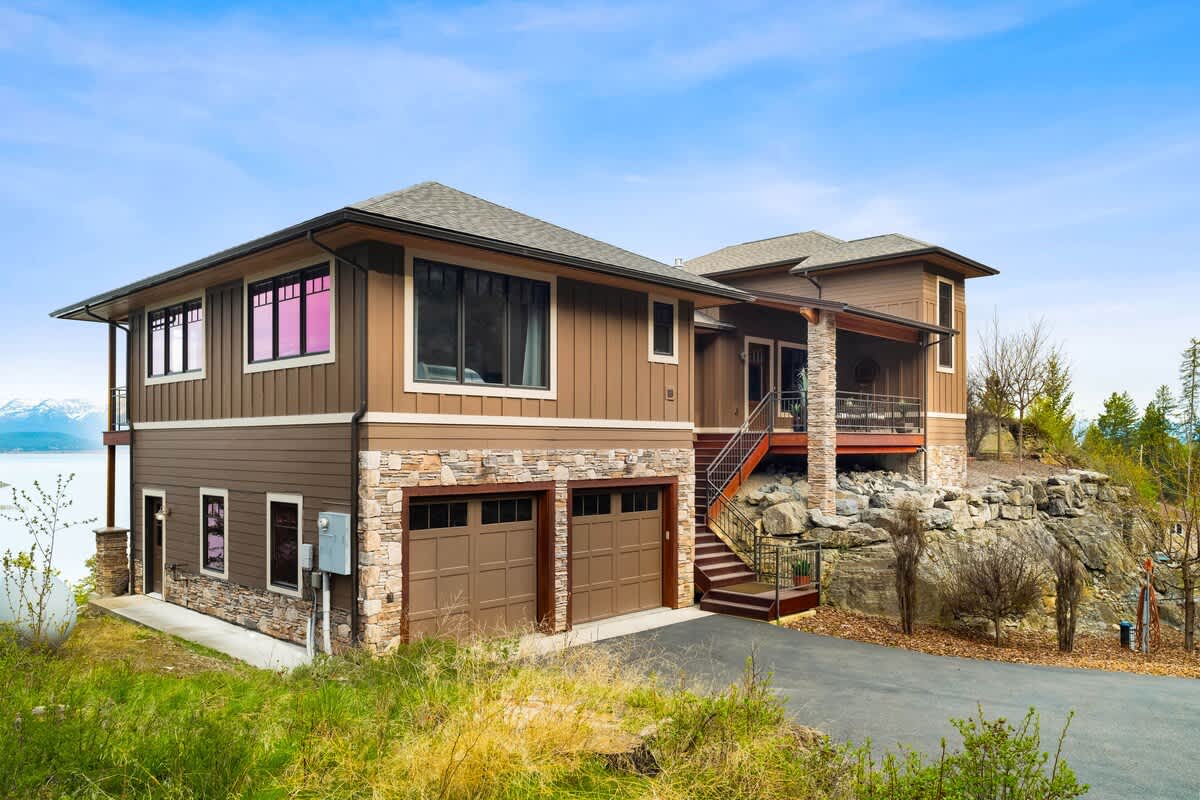 The front exterior of a modern brown house with stone accents and multiple levels, featuring a two-car garage and an elevated entrance with stairs. The house is built into a rocky hillside, offering views of the surrounding landscape.