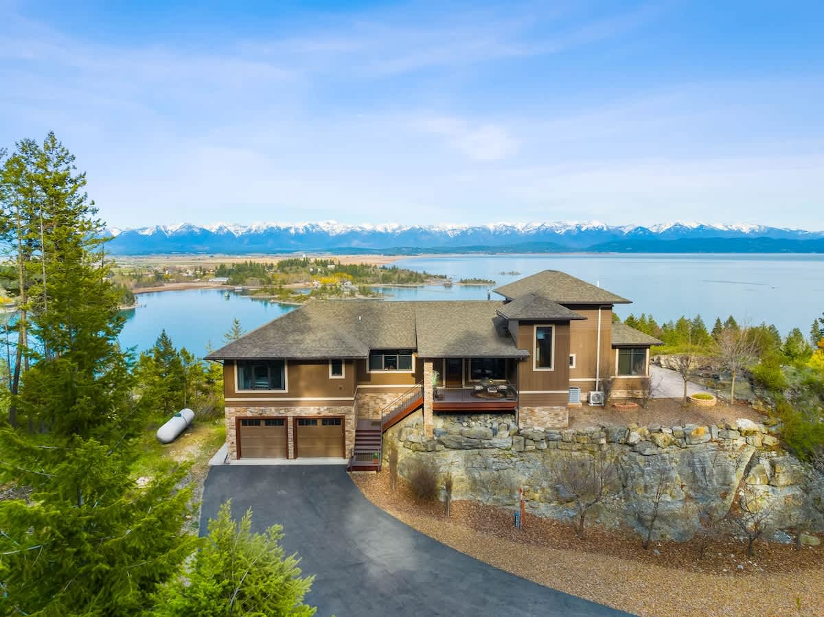 An aerial view showcases a multi-level brown house with a dark roof, perched on a rocky cliff overlooking a large blue lake and distant snow-capped mountains under a clear sky. A paved driveway leads to a two-car garage.