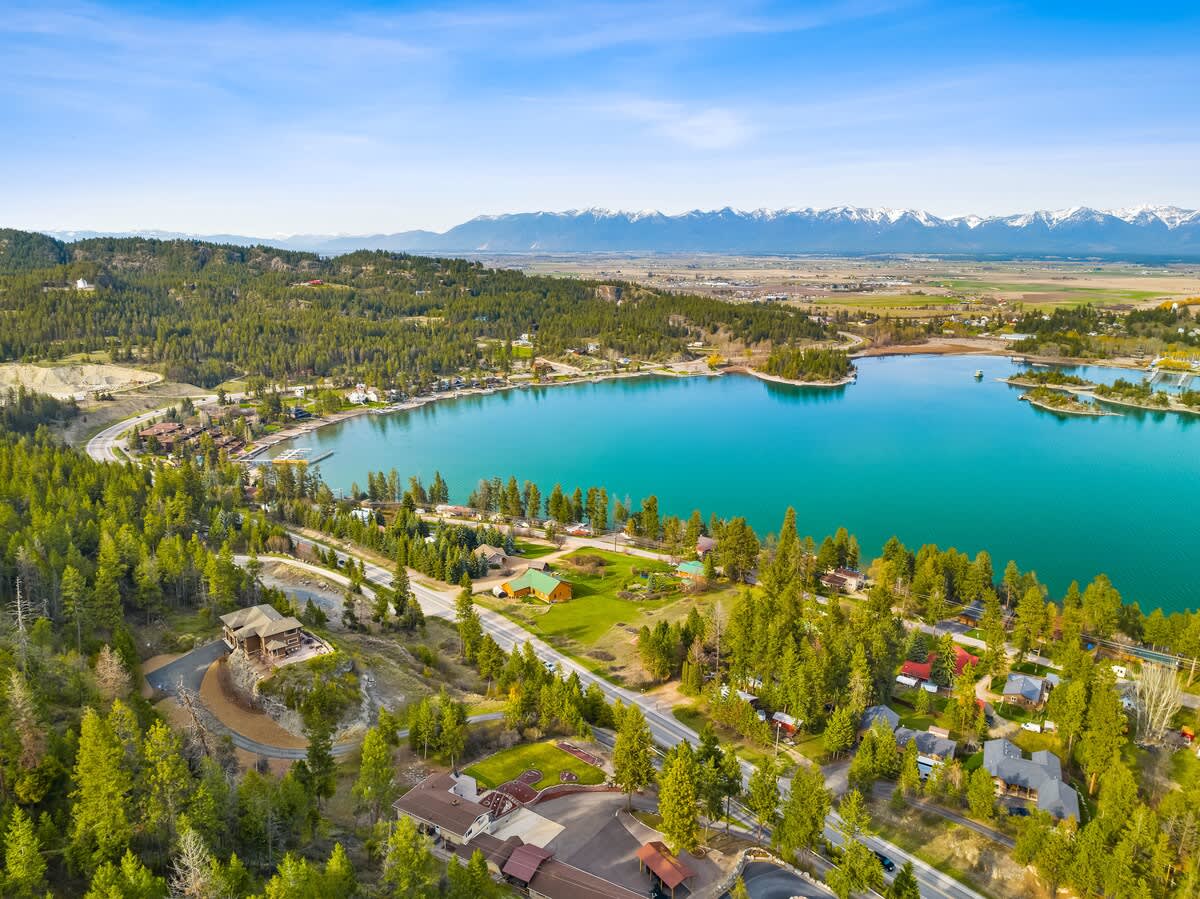 An aerial view showcasing a large lake surrounded by green forests and residential areas, with distant snow-capped mountains under a clear blue sky.