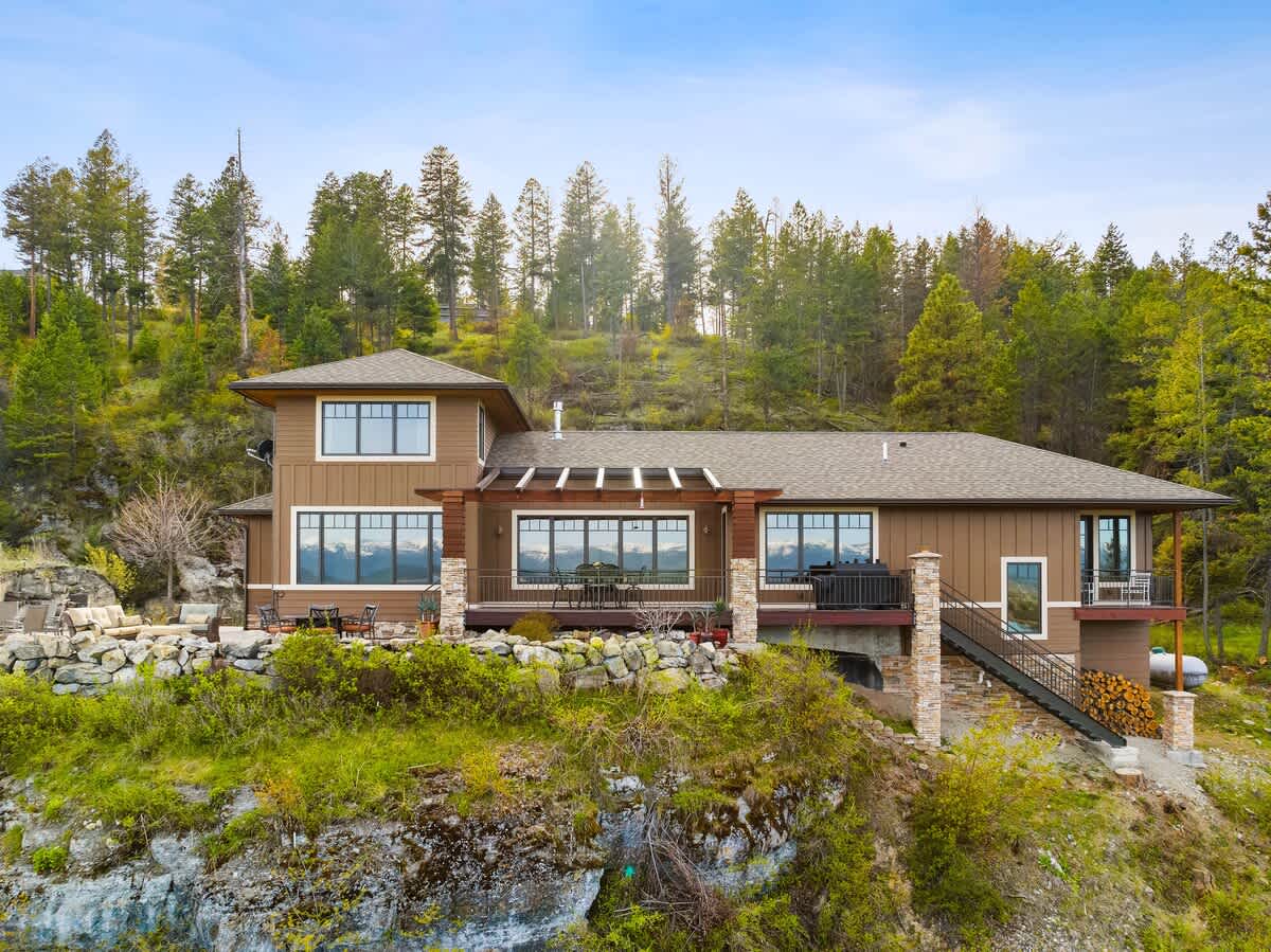 A modern brown house with stone accents and large windows, featuring an expansive deck with a wooden pergola and outdoor seating, plus a stone patio area, all nestled on a grassy cliffside with a dense forest backdrop.