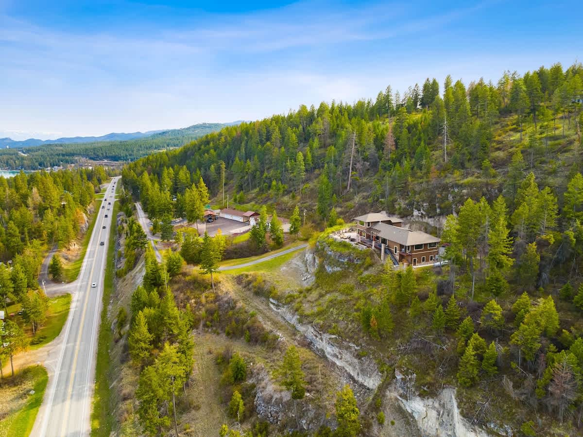An aerial perspective showing the house on a cliffside, with a winding road below and vast evergreen forests stretching into the distance, highlighting the secluded natural setting and expansive landscape.