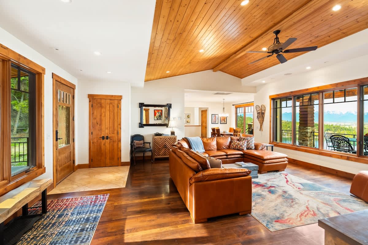 The entrance area opens into a bright living space with a vaulted wood-paneled ceiling and dark hardwood floors. A large brown leather sectional is visible, and a doorway leads to other rooms, with natural light streaming in from windows.