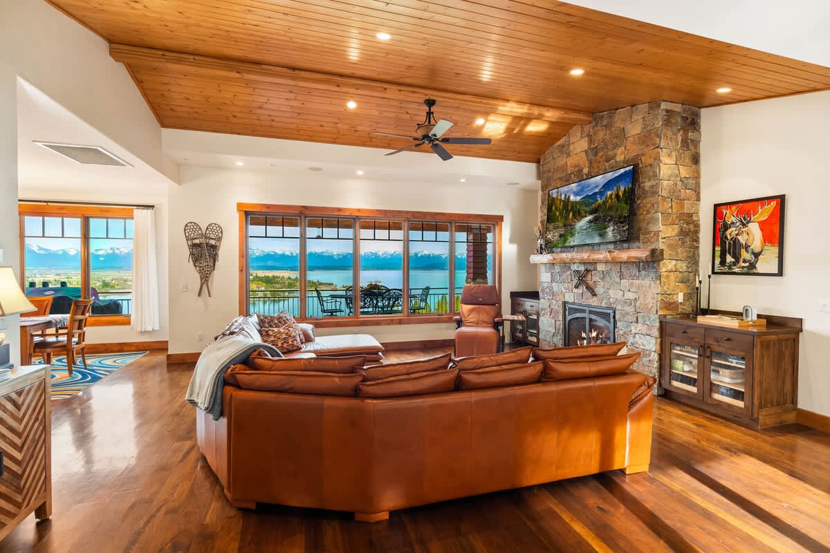 A spacious living room with a large brown leather sectional sofa facing a stone fireplace with a mounted TV. The room features a vaulted wood-paneled ceiling, hardwood floors, and expansive windows offering breathtaking lake and mountain views.