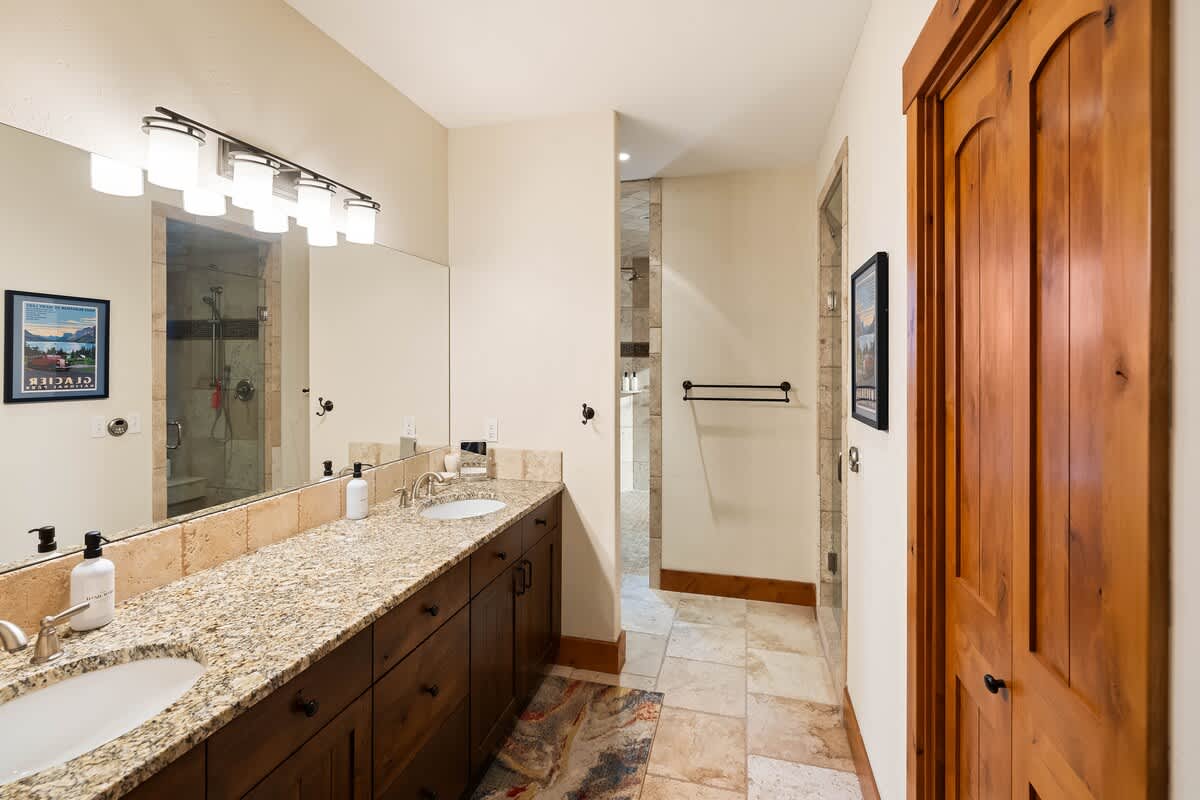 A long bathroom vanity features double sinks with granite countertops and dark wood cabinets, illuminated by a multi-bulb light fixture above a large mirror. The floor is tiled, leading towards a stone-tiled shower area.