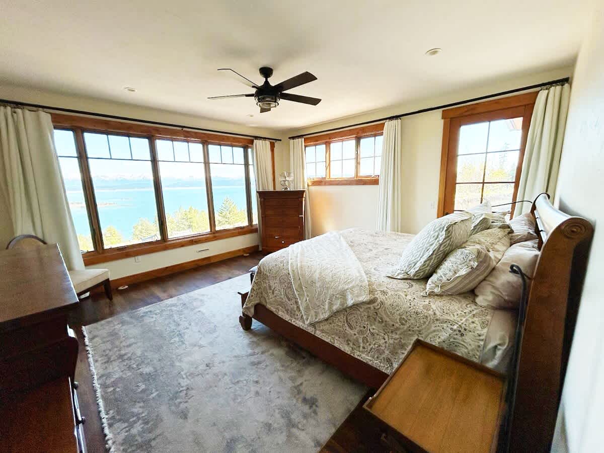 A spacious bedroom featuring a wooden sleigh bed with patterned bedding, a ceiling fan, and large windows offering panoramic lake and mountain views.