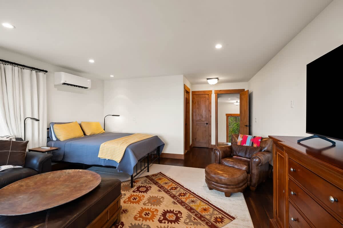 A cozy bedroom features a queen-sized bed with blue and yellow bedding, a leather armchair and ottoman, and a large flat-screen TV on a wooden dresser. The room has hardwood floors and a colorful patterned rug.