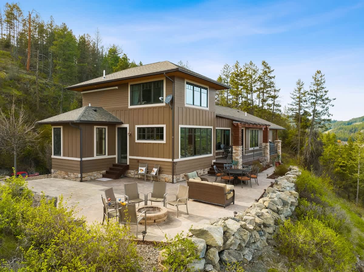 The back of the brown house features multiple windows and a large concrete patio with outdoor seating and a fire pit. The patio is bordered by a low stone wall and overlooks a scenic landscape of trees and distant views.