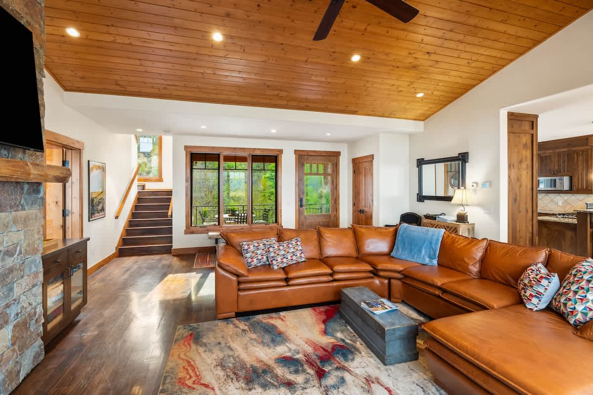 An inviting living space featuring a large brown leather sectional sofa on a patterned rug, with a stone fireplace and TV on one side. Hardwood floors extend throughout, and a wooden staircase leads to another level, all under a vaulted wood-paneled ceiling.