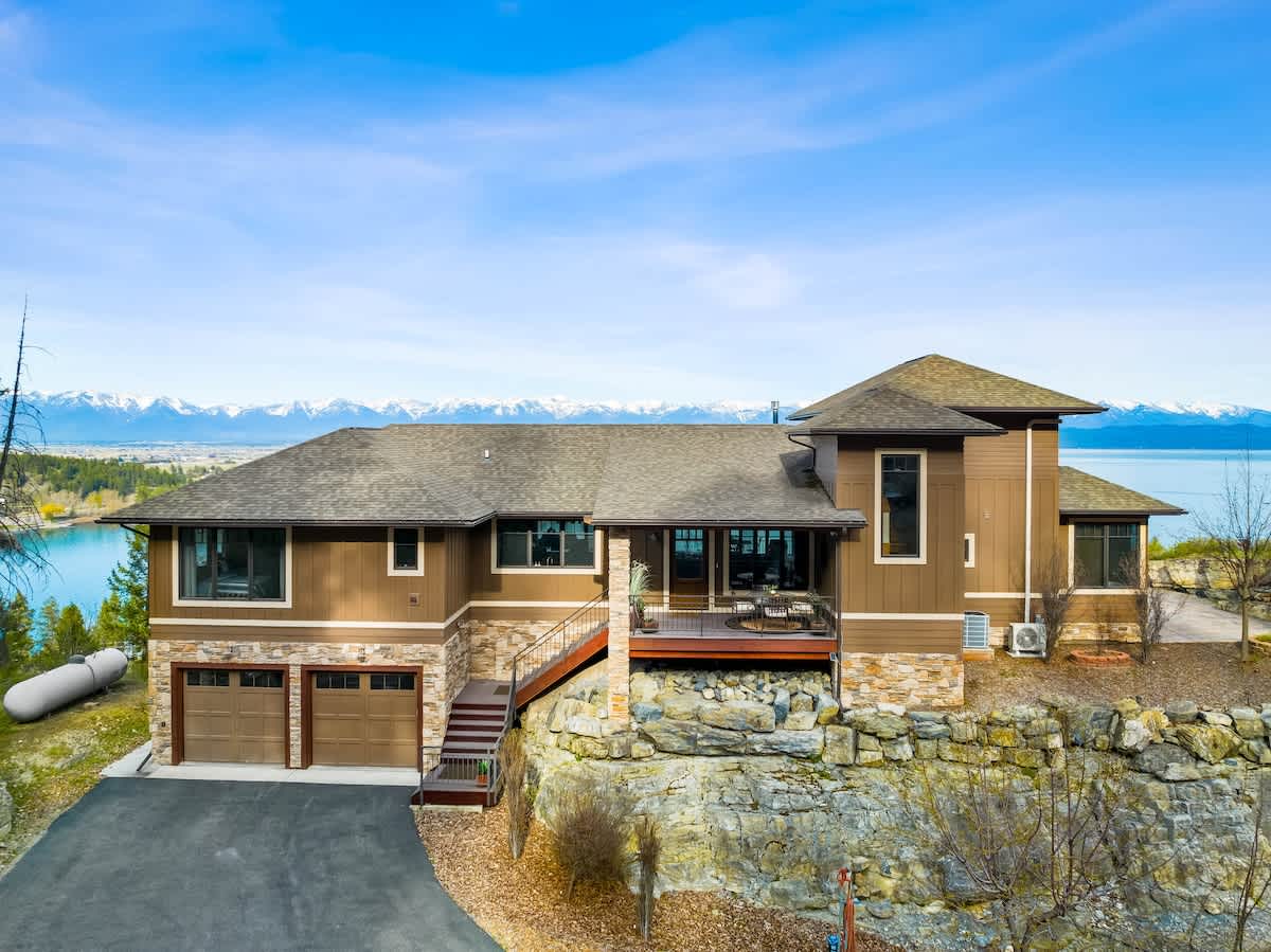 The front of a brown modern house with stone accents, featuring a three-car garage with dark wood doors and a deck with stairs leading up. A stunning blue lake and snow-capped mountain range are visible in the background.
