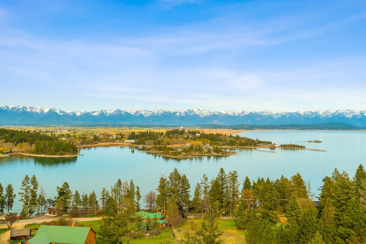 A wide aerial shot captures a serene lake with a forested peninsula and scattered homes, set against a backdrop of snow-capped mountains and a clear blue sky. The landscape emphasizes the property's expansive natural beauty.