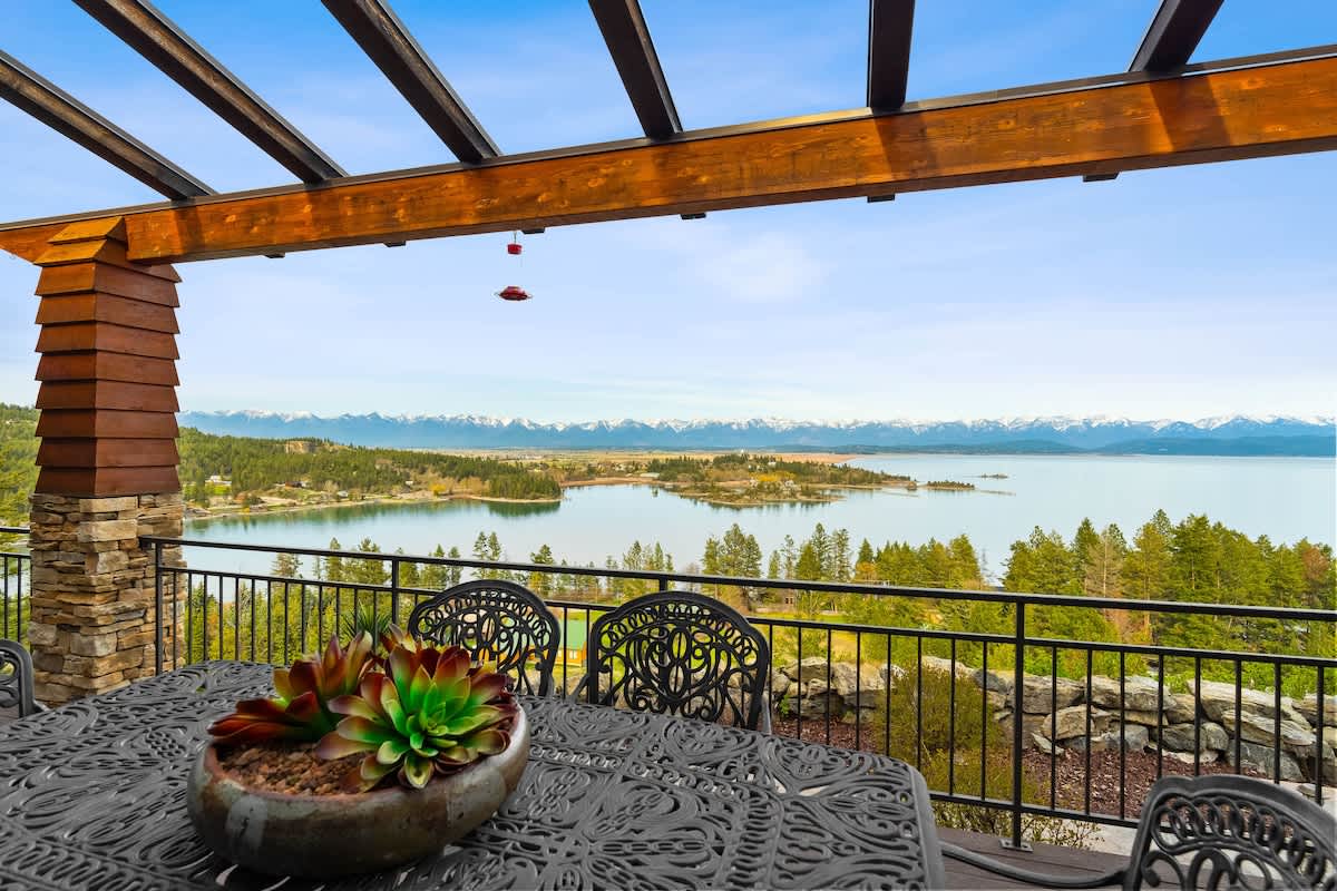A spacious outdoor deck with a dark metal dining table and chairs is covered by a wooden pergola, offering expansive views of a serene blue lake and majestic snow-capped mountains. A black metal railing secures the deck.