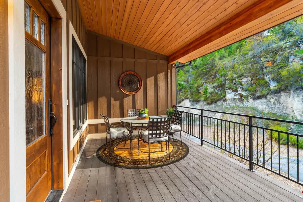 A covered outdoor deck with a round dining table and chairs, overlooking a rocky cliffside and lush greenery, featuring a decorative wall hanging.