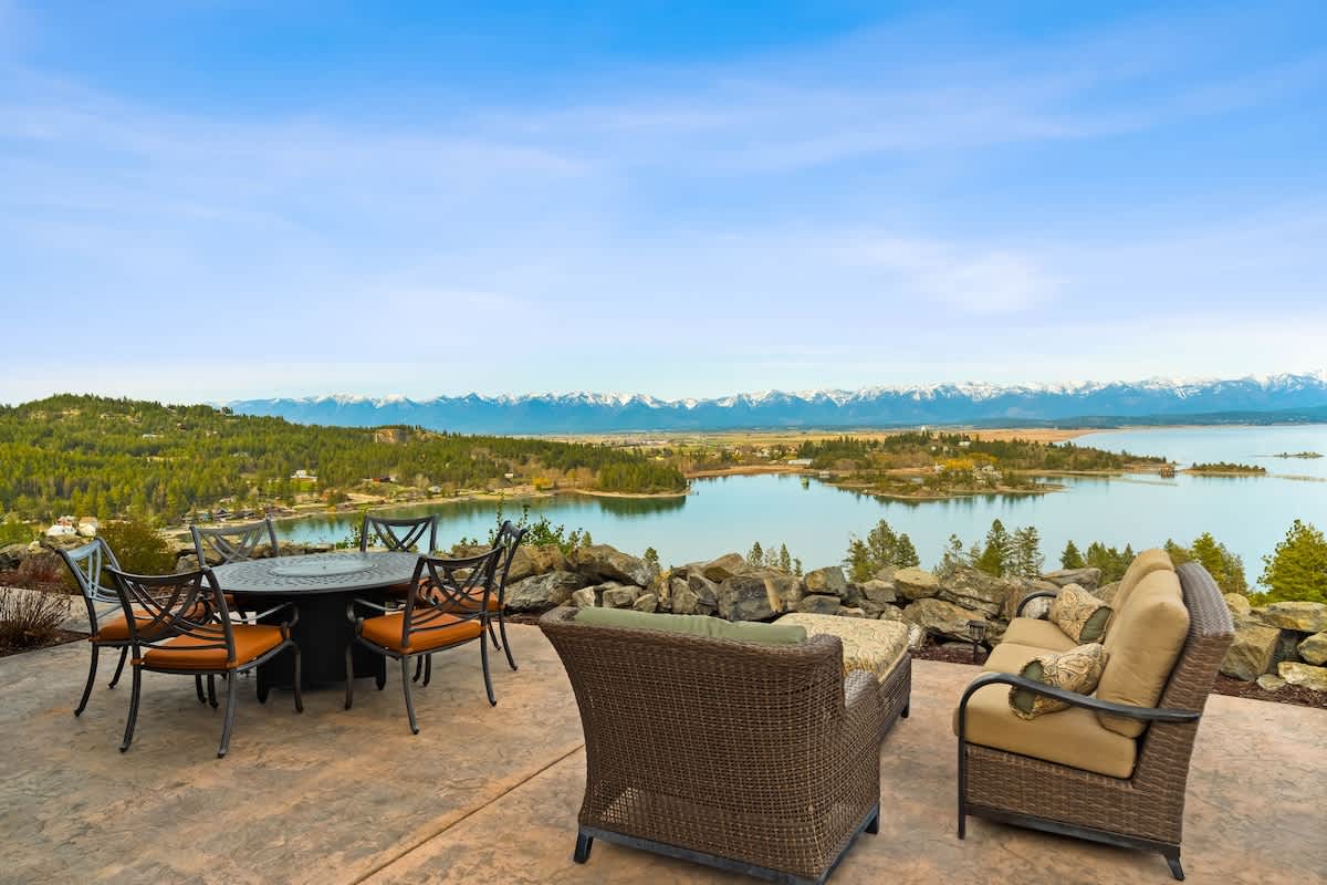 A spacious outdoor patio features a round dining table with six chairs and a wicker sofa seating area, all overlooking a large lake and distant snow-capped mountains. The patio is paved and surrounded by natural rock landscaping.