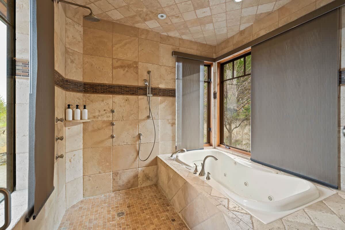 A spacious bathroom features a large walk-in shower with stone tiles and a rain showerhead, alongside a jetted bathtub positioned by a window with privacy blinds. The room has warm-toned tiled floors and recessed lighting.