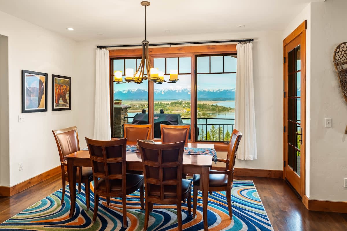 A bright dining room featuring a rectangular wooden table with six leather chairs, set on a vibrant blue, orange, and green patterned rug. Large windows frame stunning views of a lake and snow-capped mountains, with white curtains and a rustic chandelier.