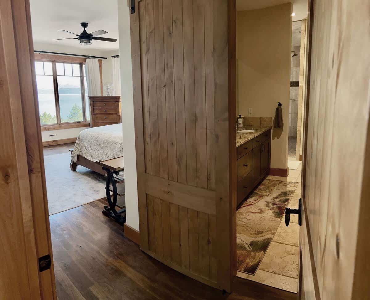 A rustic wooden barn door slides open to reveal a bathroom vanity with a sink, tiled backsplash, and a patterned rug, with the bedroom visible beyond.