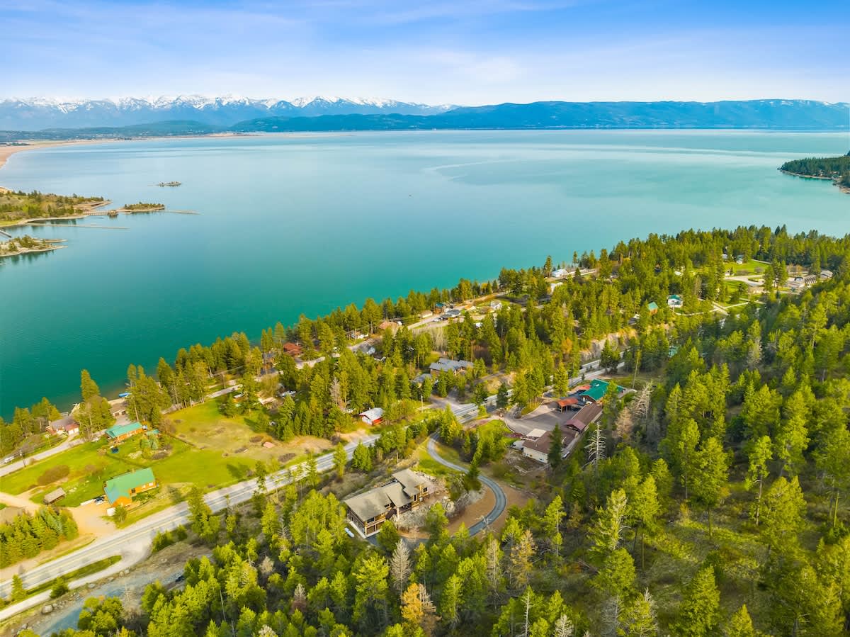 An aerial view showcasing a large house nestled among lush green trees on a hillside, overlooking a vast, turquoise lake with distant snow-capped mountains. A winding road is visible, connecting the property to the scenic landscape.