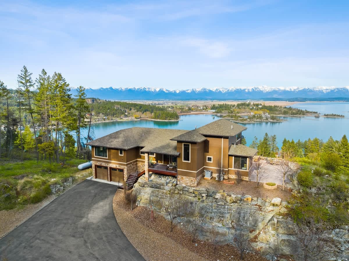 A wide aerial shot of the brown house with its three-car garage, situated on a rocky outcrop, offering panoramic views of a large blue lake and majestic snow-capped mountains under a bright, clear sky.