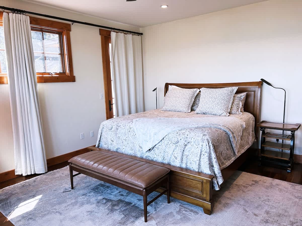 A cozy bedroom with a wooden sleigh bed, patterned bedding, a leather bench at the foot, and a tall lamp beside a dark wood nightstand.