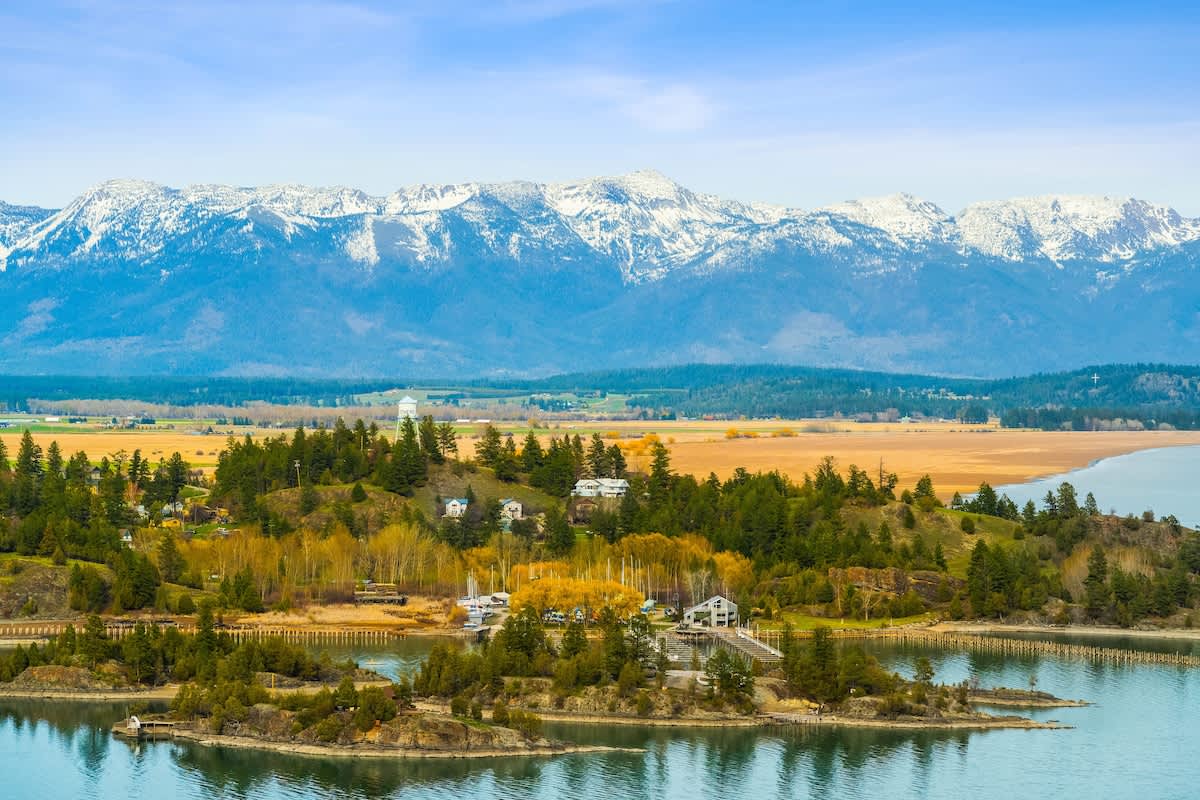 An aerial view showcases a lake with small islands and a forested shoreline, backed by a vast plain and snow-capped mountains under a blue sky. The scene highlights the property's scenic natural surroundings.