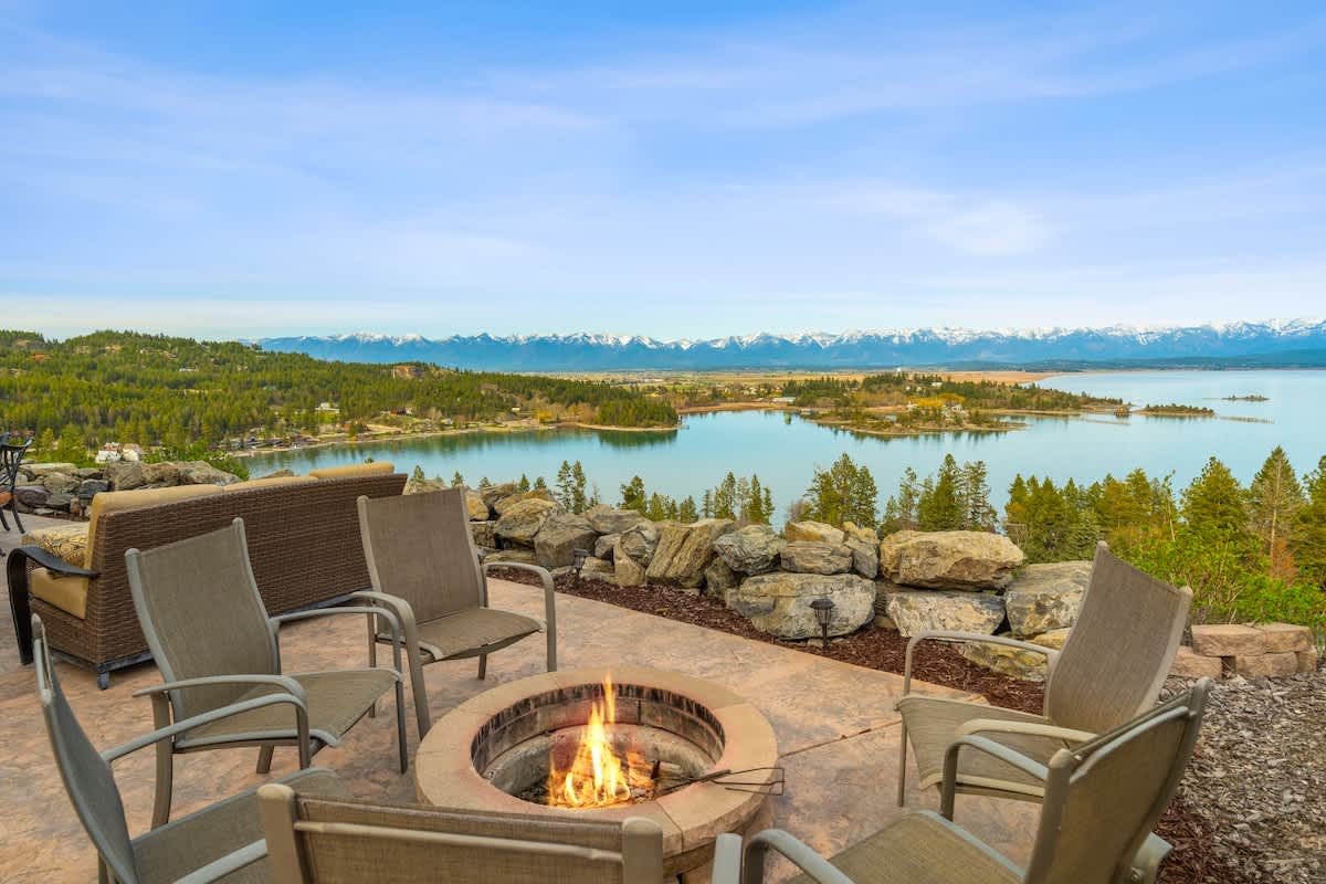 An outdoor fire pit area features a circular stone fire pit surrounded by several chairs on a concrete patio, all set against a stunning backdrop of a blue lake and snow-capped mountains. Low stone walls and greenery frame the space.