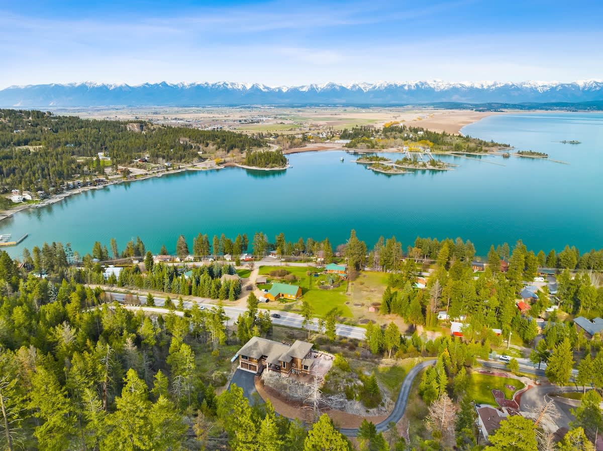 A wide aerial shot captures the brown house nestled among green trees on a hillside, overlooking a large, calm blue lake with several islands and distant snow-capped mountains. A winding road is visible nearby.