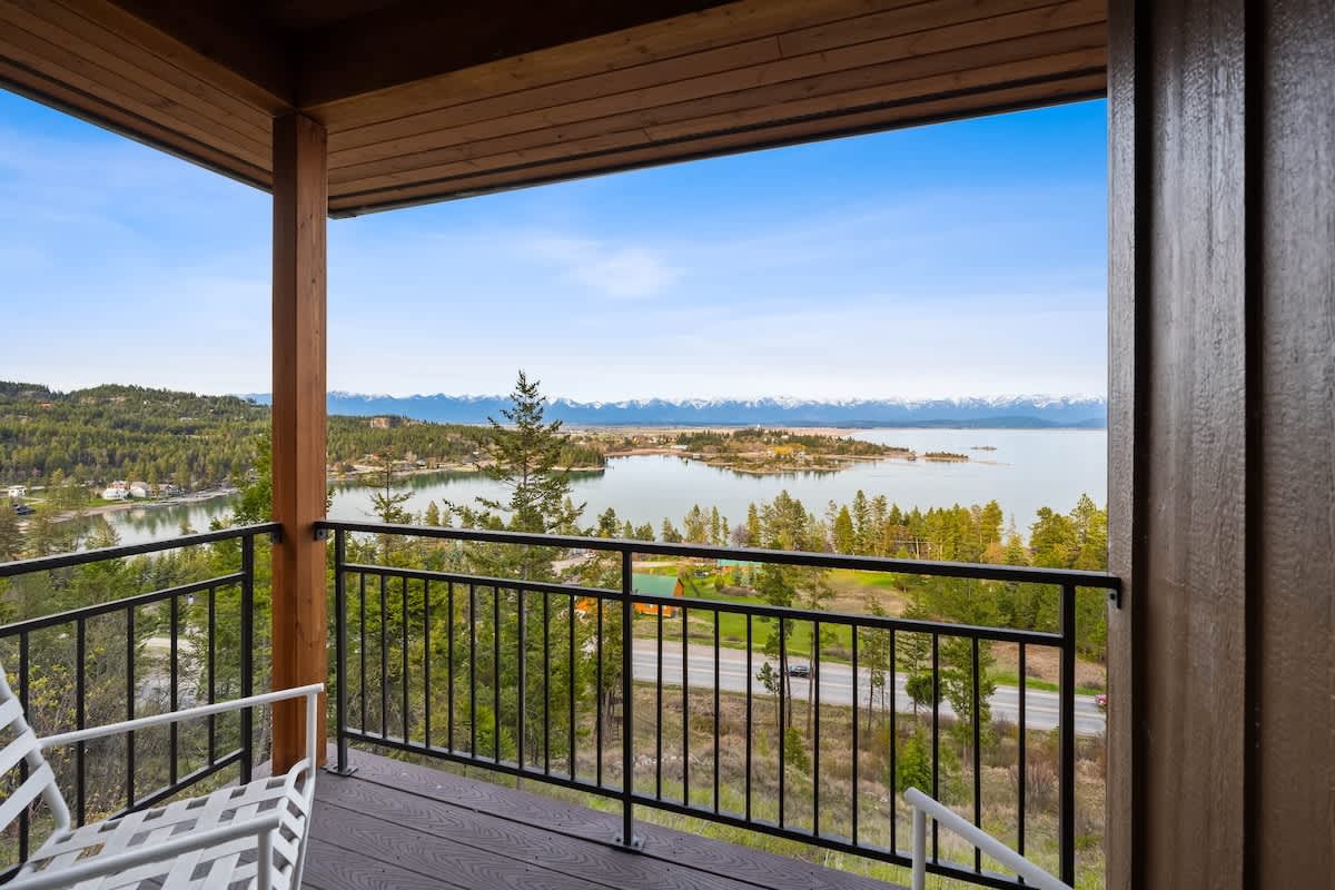 A private balcony with black metal railings offers expansive views of a lake, forested hills, and distant snow-capped mountains under a bright blue sky. A white chair provides a spot for relaxation.