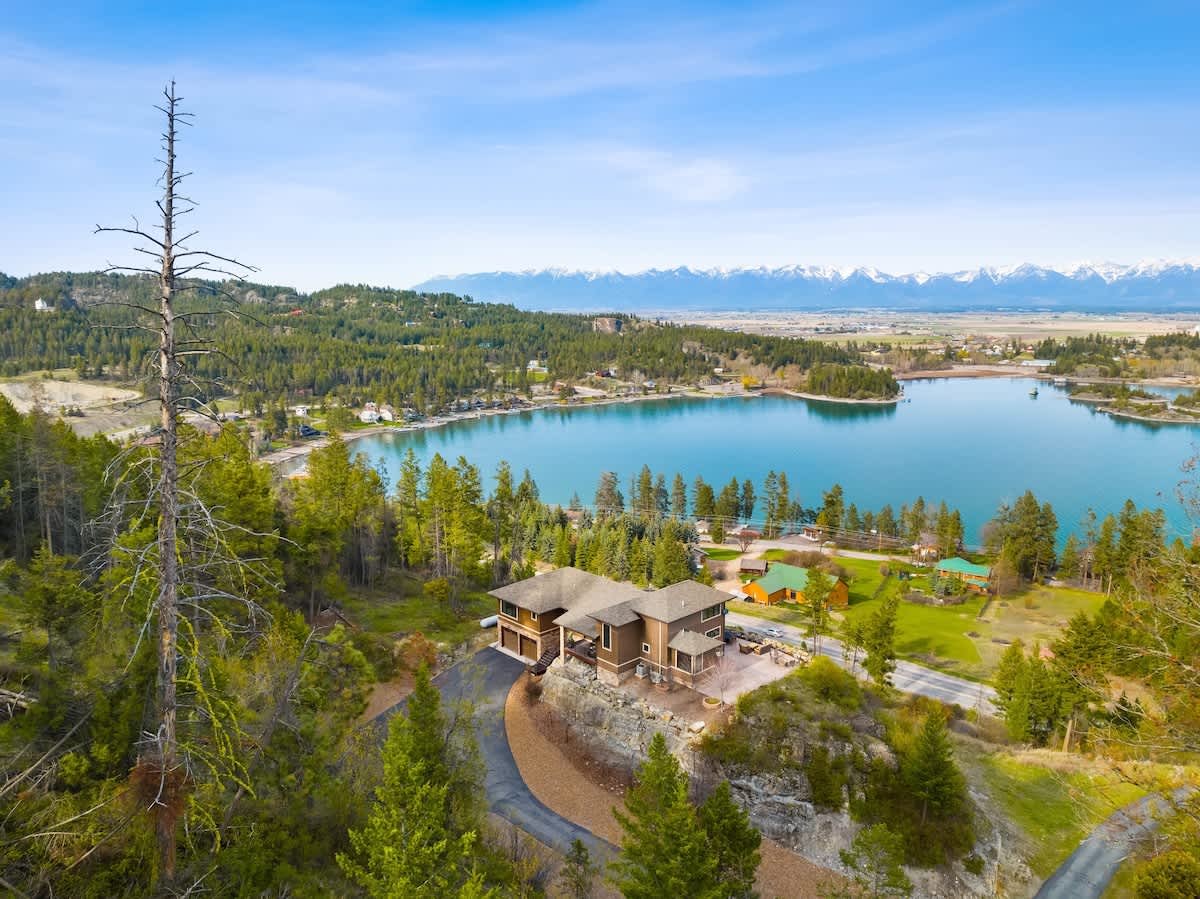 A high aerial view capturing the brown house perched on a green hillside, overlooking a serene blue lake and distant snow-capped mountains under a clear sky, surrounded by dense evergreen forest.