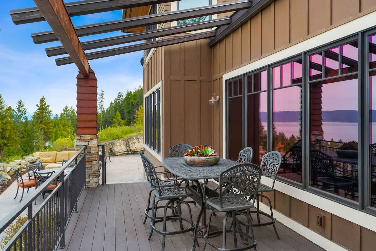 A wide view of the outdoor deck shows a dark metal dining set under a wooden pergola, with the brown house exterior visible through large windows. The deck extends to another patio area with additional seating, all overlooking the scenic landscape.