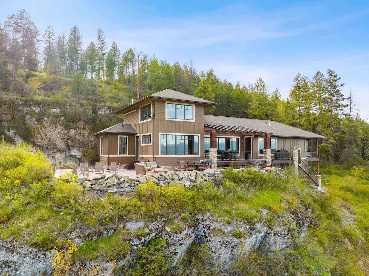 An elevated view of the brown-sided house, showcasing its extensive deck with a wooden pergola and a stone patio area, complete with outdoor furniture, set against a lush green hillside and a backdrop of tall pine trees.