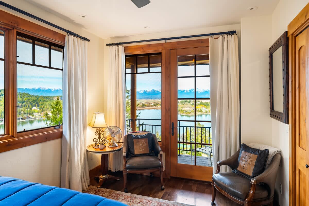 A cozy corner in the bedroom with two leather armchairs, a small round table with a lamp, and a glass door opening to a balcony with scenic lake and mountain views.