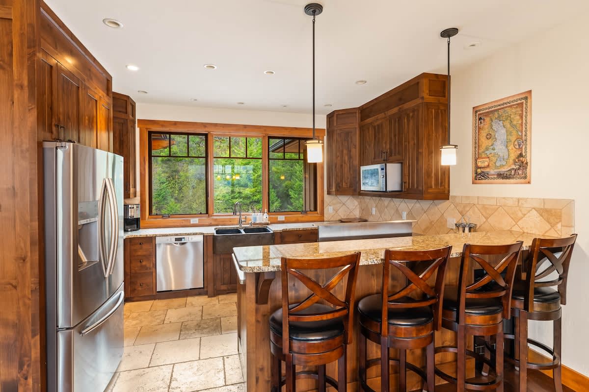 A well-appointed kitchen featuring dark wood cabinetry, granite countertops, and stainless steel appliances, including a large refrigerator and dishwasher. A breakfast bar with four high-backed stools provides casual seating, with a large window looking out to lush greenery.