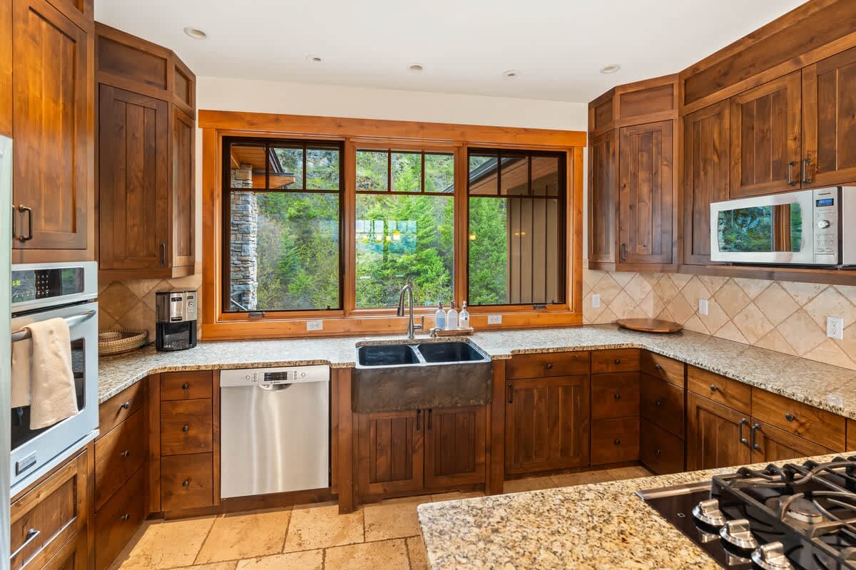A detailed view of the kitchen's U-shaped layout, showcasing dark wood cabinets, speckled granite countertops, and a large farmhouse-style sink. Stainless steel appliances like a dishwasher and built-in microwave are visible, with a window offering a view of trees.