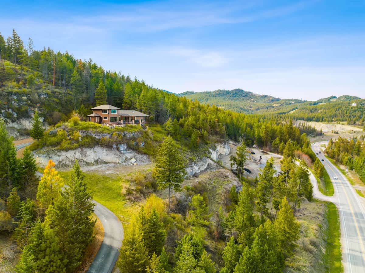 An aerial perspective shows the brown house situated on a forested hillside, with a winding paved driveway leading up to it. A road runs along the base of the hill, and distant green hills are visible under a clear sky.