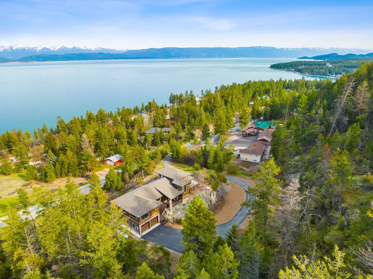 An aerial perspective highlighting the property nestled among green trees on a hillside, overlooking a vast lake with distant mountains under a bright sky.