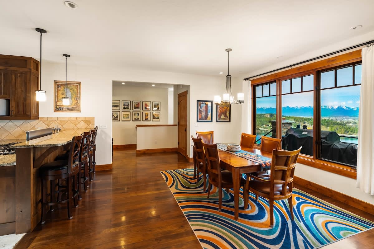 An open-concept dining area with a wooden table and chairs on a vibrant rug, connected to a kitchen with a breakfast bar. Large windows offer expansive views of the lake and mountains, creating a bright and inviting space.