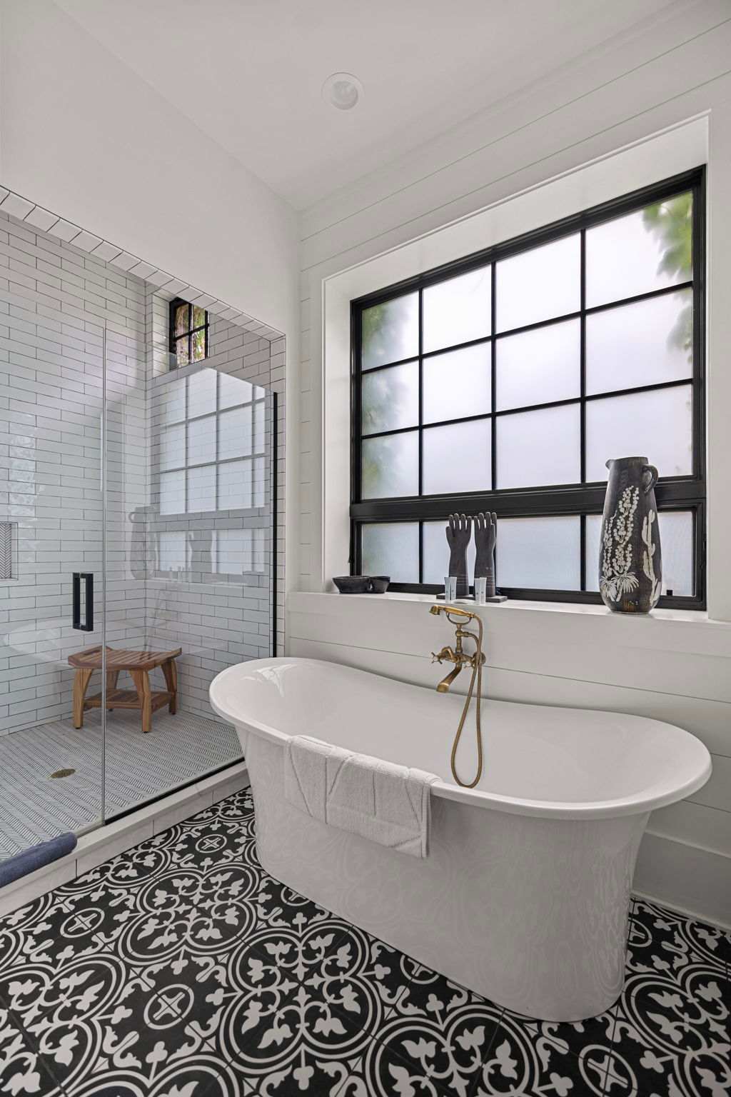 Modern bathroom with a freestanding white soaking tub and brass fixtures, set against white shiplap walls. A large black-framed window with frosted glass provides privacy, while the floor features striking black and white patterned tiles.