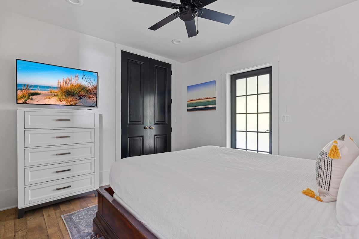 A bright bedroom features a bed with crisp white bedding, a white dresser topped with a flat-screen TV, and a black ceiling fan. Black paneled doors and a black-framed glass door add contrast to the white walls.