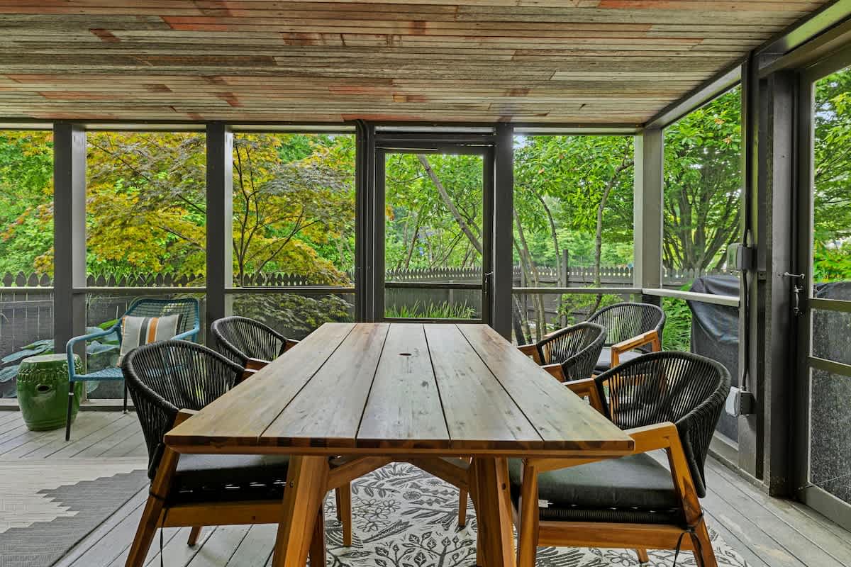 Screened-in porch with a long wooden dining table surrounded by black woven chairs, set on a patterned rug, offering an outdoor dining experience with garden views.