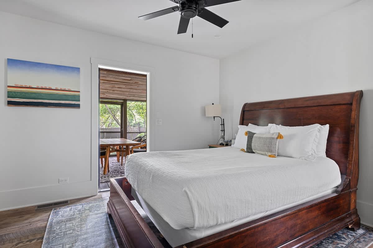 This bedroom features a classic wooden sleigh bed with white bedding and decorative pillows, facing a doorway that opens to a screened outdoor area. A black ceiling fan is visible above, and a colorful abstract painting hangs on the wall.