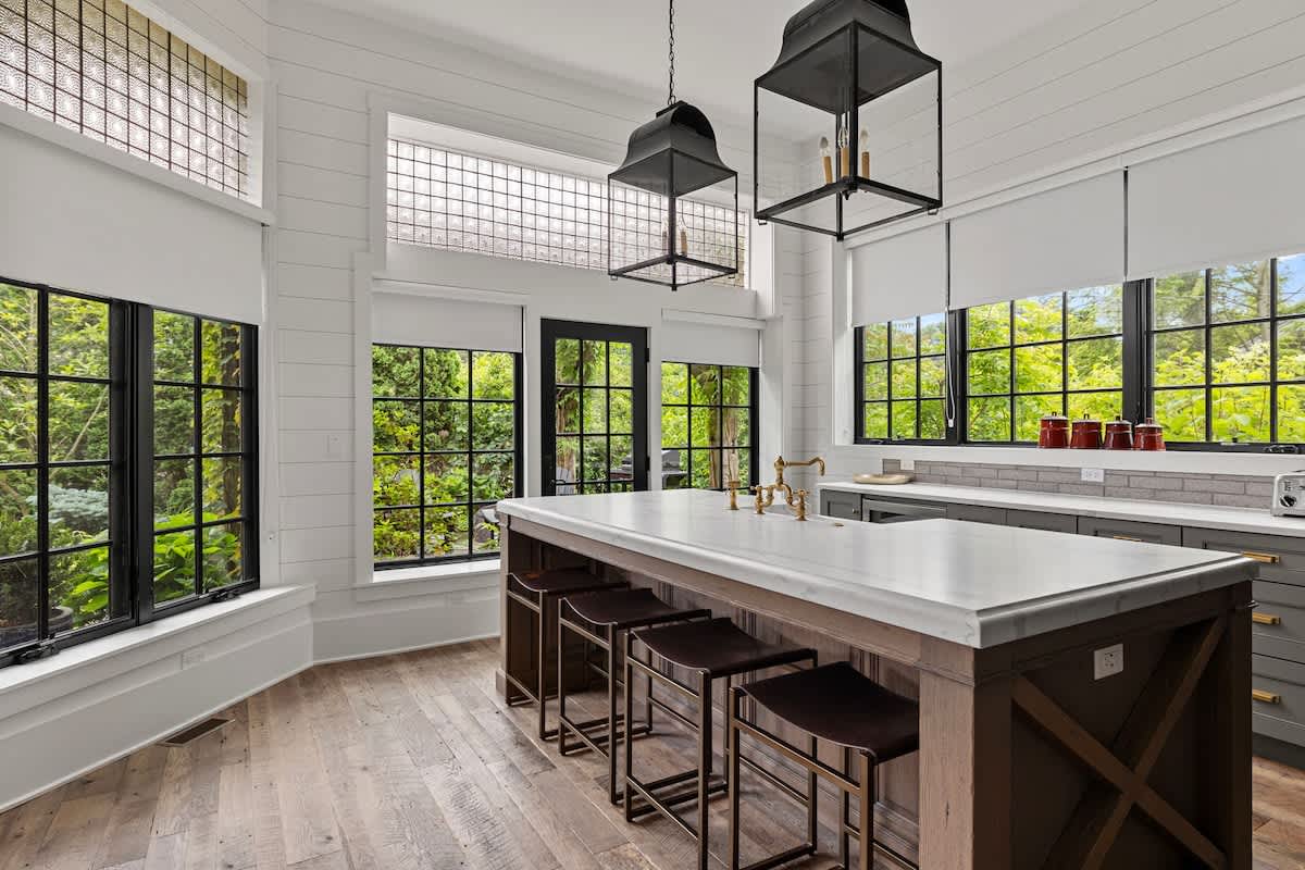 Bright kitchen featuring a large white marble island with a dark wood base and four dark leather bar stools. Black-framed windows offer views of lush greenery, complemented by black lantern pendant lights and white shiplap walls.