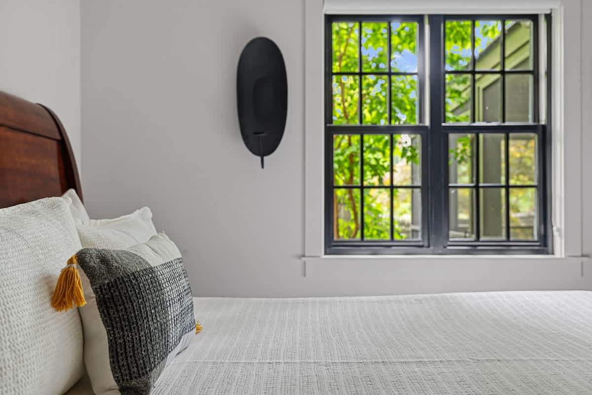 A cozy bedroom with a dark wood headboard and white bedding, featuring a large window with black frames and multiple panes, offering a view of lush green trees outside. A black wall sconce adds a modern touch.