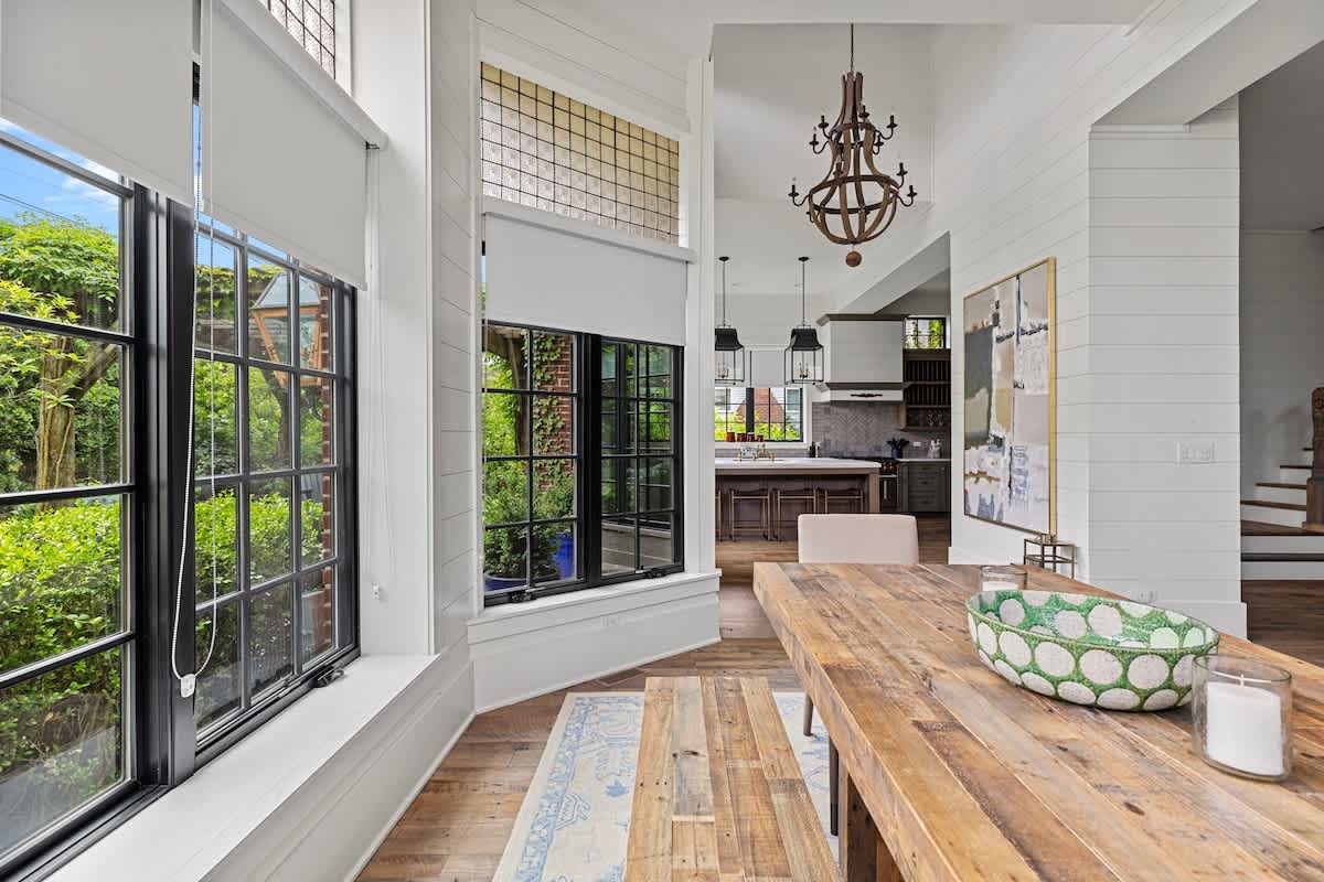 A long rustic wooden dining table with a matching bench is centered in a bright room, featuring a green patterned bowl and candles. Large black-framed windows provide ample natural light and views of the greenery, with high ceilings and a chandelier in the adjacent space.