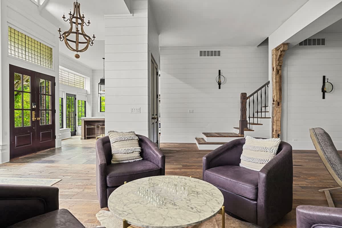 An elegant living area with dark purple swivel chairs and a marble coffee table, featuring shiplap walls, hardwood floors, and a view towards the main entrance and staircase.