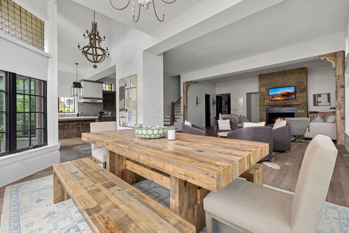 Open-concept dining area featuring a large rustic wooden dining table with matching benches and upholstered chairs, set on a light blue patterned rug. The space flows into a living room and a modern kitchen.