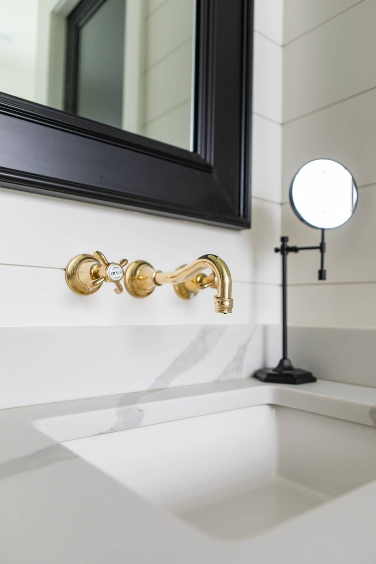 A close-up of the bathroom vanity, highlighting a wall-mounted gold faucet against white shiplap walls and a white marble-patterned countertop with an undermount sink. A black-framed mirror is visible above.