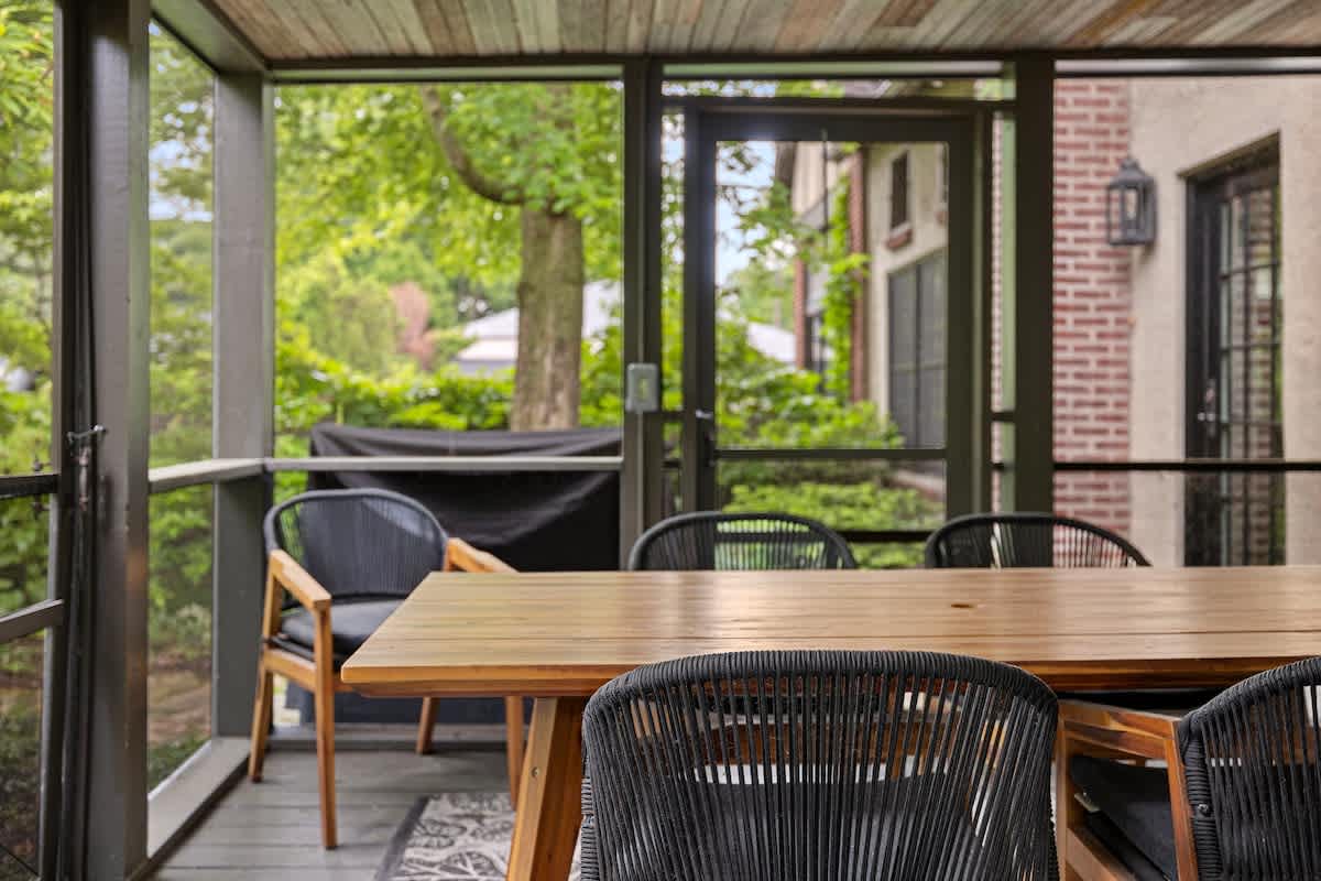 Screened-in porch featuring a wooden dining table and black woven chairs, set on a patterned rug. Lush green trees are visible through the screens, with a brick exterior wall in the background.