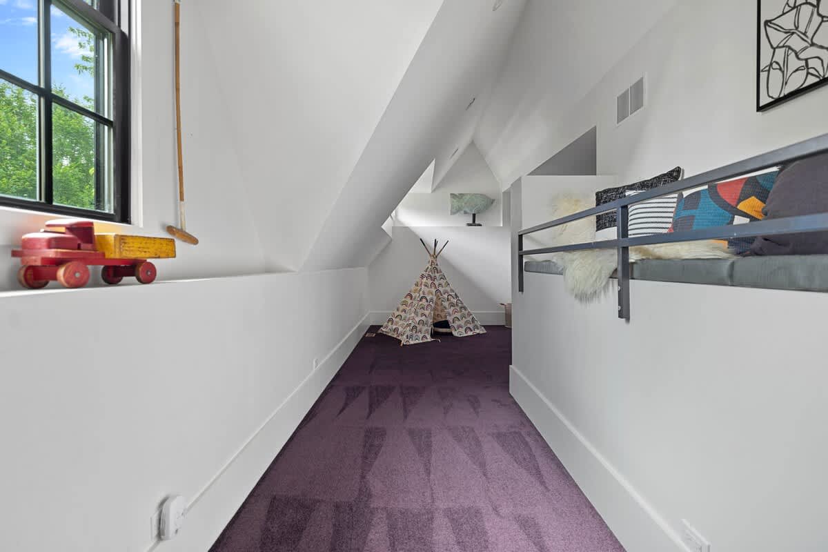 Cozy attic play area with a small teepee, a window seat with cushions, and purple carpet, featuring a sloped ceiling and a large window overlooking greenery.