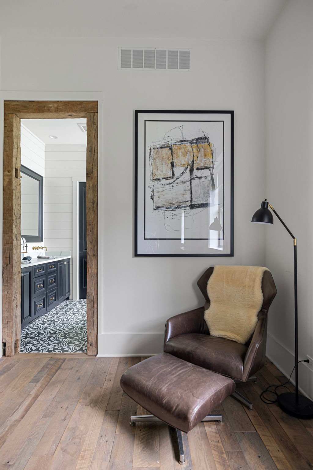 A comfortable sitting area with a brown leather armchair and ottoman, accented with a sheepskin throw, next to a modern black floor lamp. A large abstract artwork hangs on the white wall, and a rustic wooden doorway leads to a bathroom with patterned tiles.